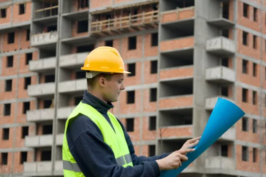 Um homem com capacete, segurando um papel azul, em um cenário que sugere trabalho de construção.