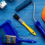 Ferramentas para pintura dispostas em uma mesa azul, temos capacete, pincel, rolo de pintura