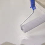 Imagem de um rolo de pintura aplicando tinta branca no chão de cimento.