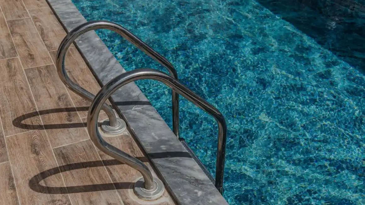 Imagem da borda de uma piscina com uma escada metálica instalada, com piso de cerâmica ao redor