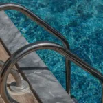 Imagem da borda de uma piscina com uma escada metálica instalada, com piso de cerâmica ao redor