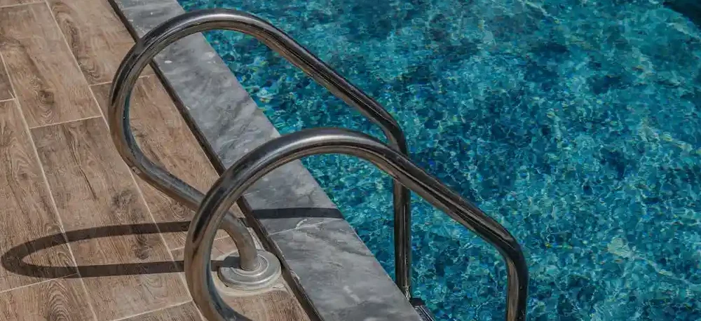 Imagem da borda de uma piscina com uma escada metálica instalada, com piso de cerâmica ao redor