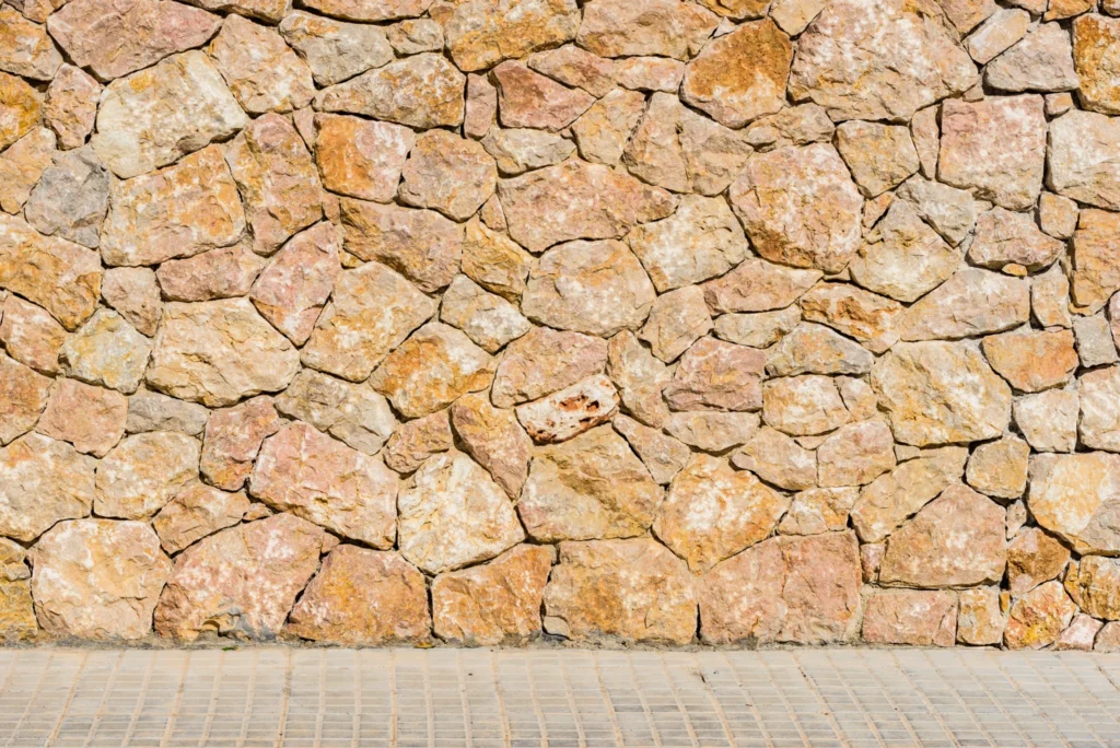 Parede de pedras irregulares em tons terrosos junto a calçada de blocos claros