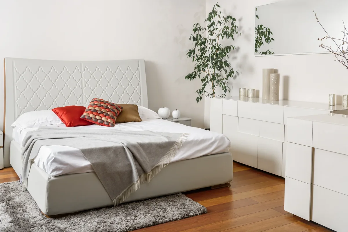 Quarto de casal branco com cabeceira estofada e almofadas vermelha e laranja como cores de destaque